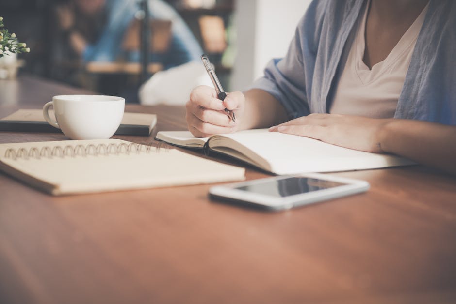 A woman writes in a notebook at a café table with a coffee and smartphone nearby